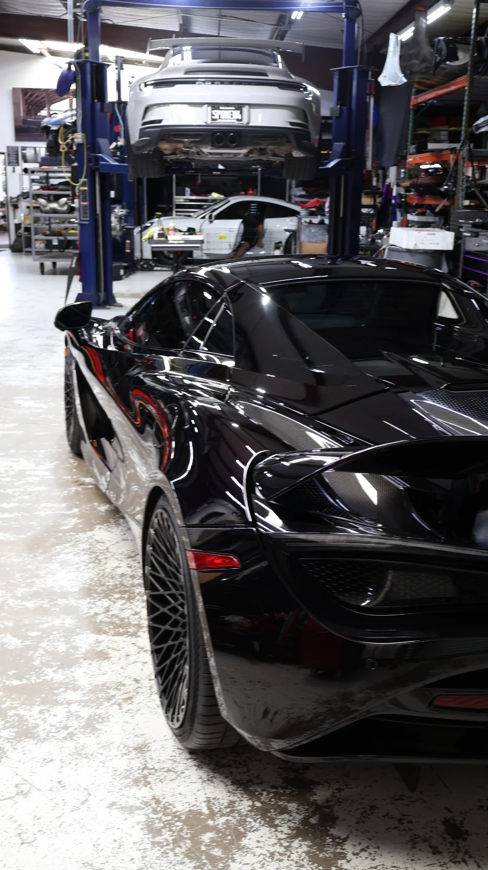 A black McLaren supercar parked in a performance tuning shop with a white Porsche on a lift in the background.