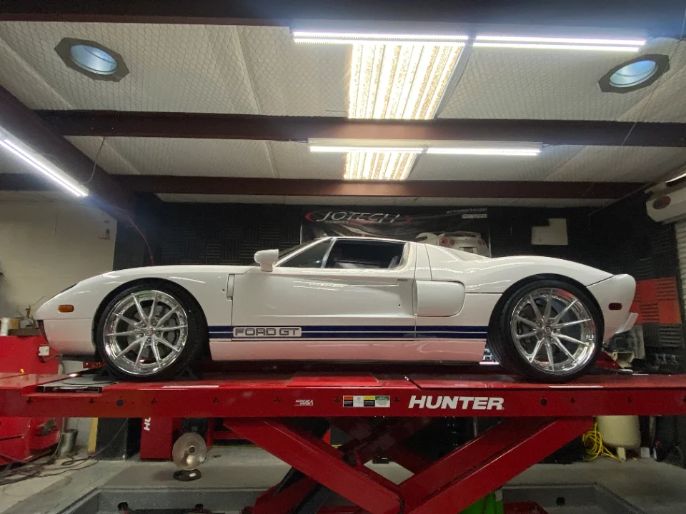 A white Ford GT with blue racing stripes sits elevated on a red car lift inside a Jotech Motorsports shop.
