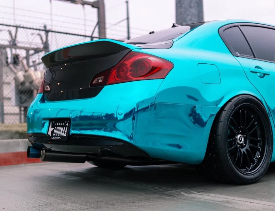 Rear view of a teal chrome-wrapped Infiniti G37 sedan with an aftermarket spoiler, black wheels, and custom exhaust.