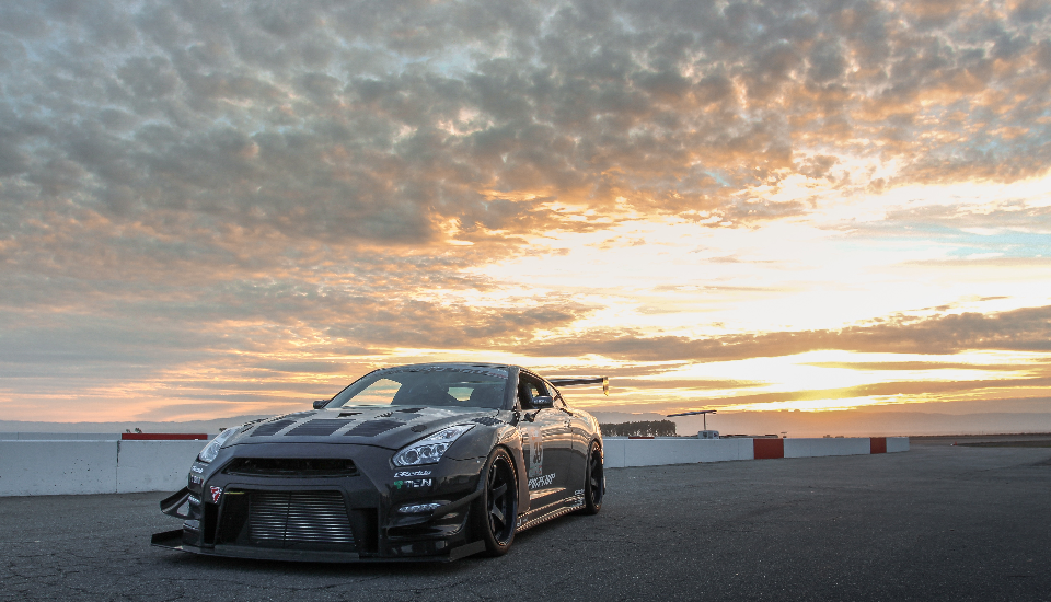A black widebody Nissan GT-R with a large front splitter and rear wing parked on a track at sunset.
