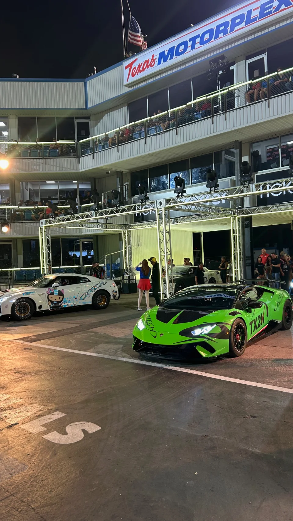 A green Lamborghini Huracan and a white Nissan GT-R with anime livery in the staging lanes at Texas Motorplex during a night event.