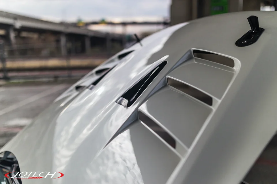 Close-up of a white performance car's hood featuring aggressive vents and a black hood pin, with a blurred outdoor background.