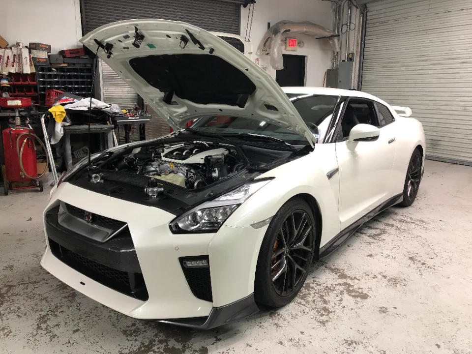 A white Nissan GT-R with its hood open, revealing the engine bay, parked inside a Jotech Motorsports shop.