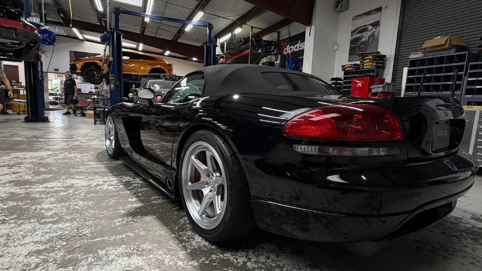 A black Dodge Viper convertible parked on a concrete floor in a performance tuning shop, with other cars on lifts in the background.