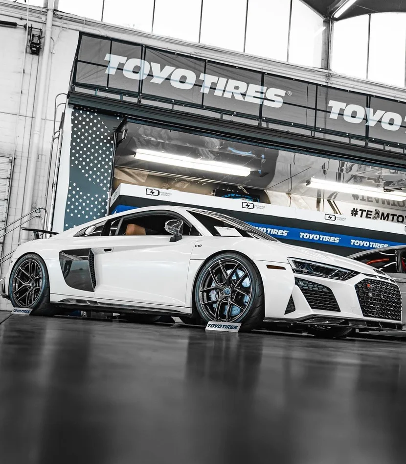 White Audi R8 with aftermarket wheels and Toyo Tires parked indoors at an event.