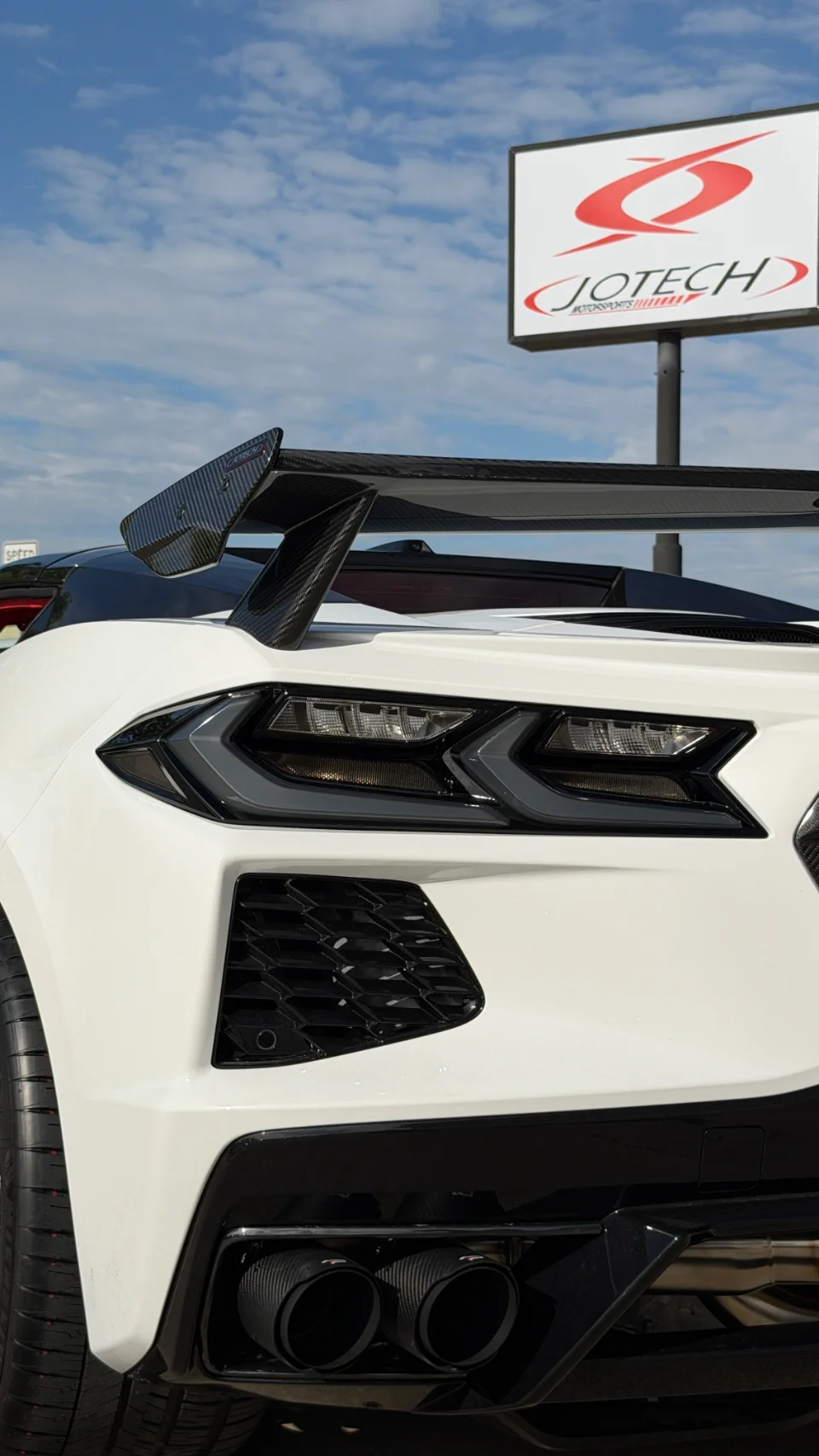 Rear view of a white C8 Corvette with a large carbon fiber wing and aftermarket exhaust tips, parked at Jotech Motorsports.
