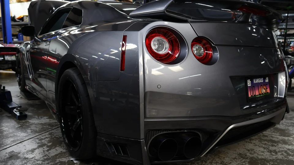Rear view of a grey Nissan GT-R with an open hood in a performance shop, showcasing its exhaust and tail lights.