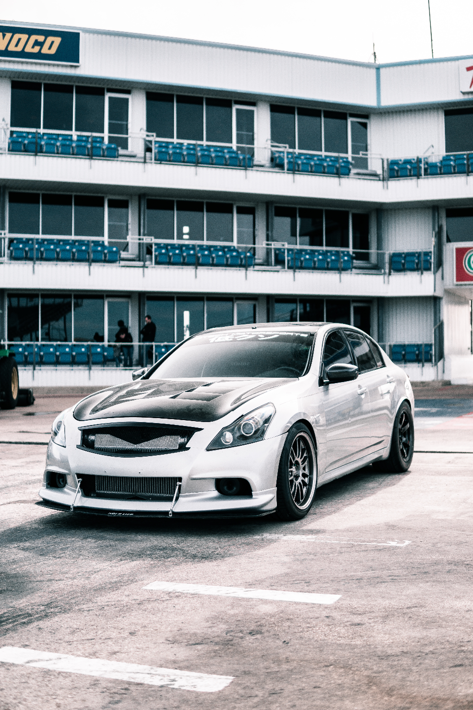 A silver Infiniti G37 sedan with a carbon fiber hood and aftermarket wheels parked at a drag strip with grandstands in the background.