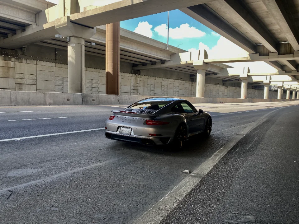 A silver Porsche 911 Turbo S parked on a highway under a concrete overpass with a blue sky visible above.