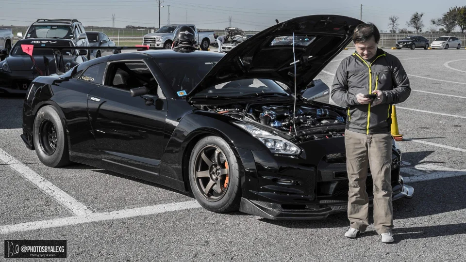 A black Nissan GT-R with its hood open and drag tires, parked at a track day event, with a man looking at his phone.