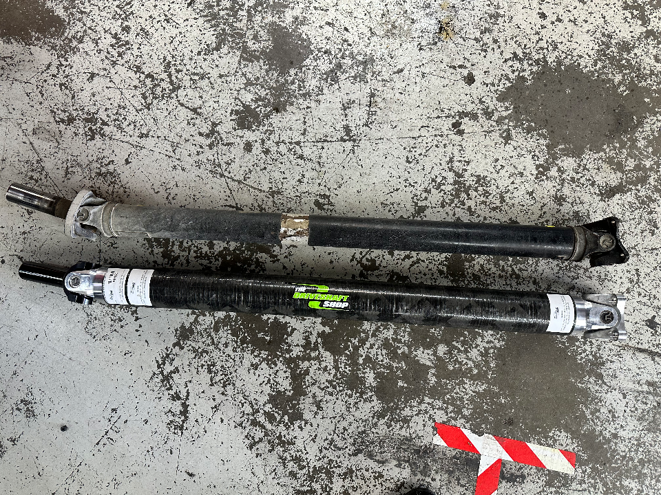Comparison of a stock driveshaft and a carbon fiber aftermarket driveshaft on a shop floor.