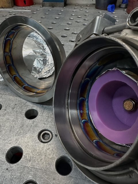 Close-up of custom fabricated stainless steel V-band exhaust components with visible welds on a metal workbench in a shop.