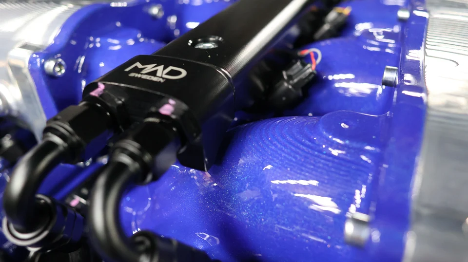 A close-up of a black MAD Sweden fuel rail and braided lines on a custom blue intake manifold in an engine bay.
