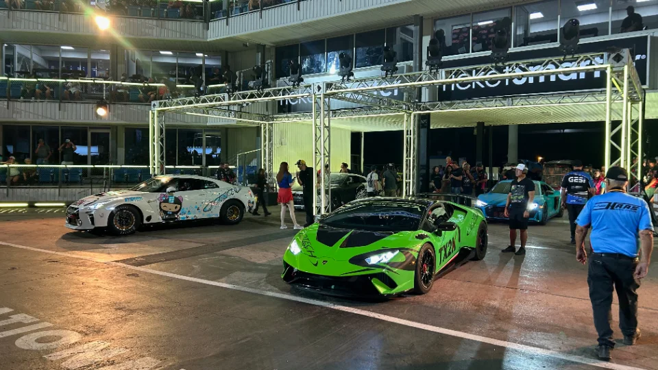 A white Nissan GT-R and a green Lamborghini Huracan at a night drag racing event, surrounded by spectators and event lighting.