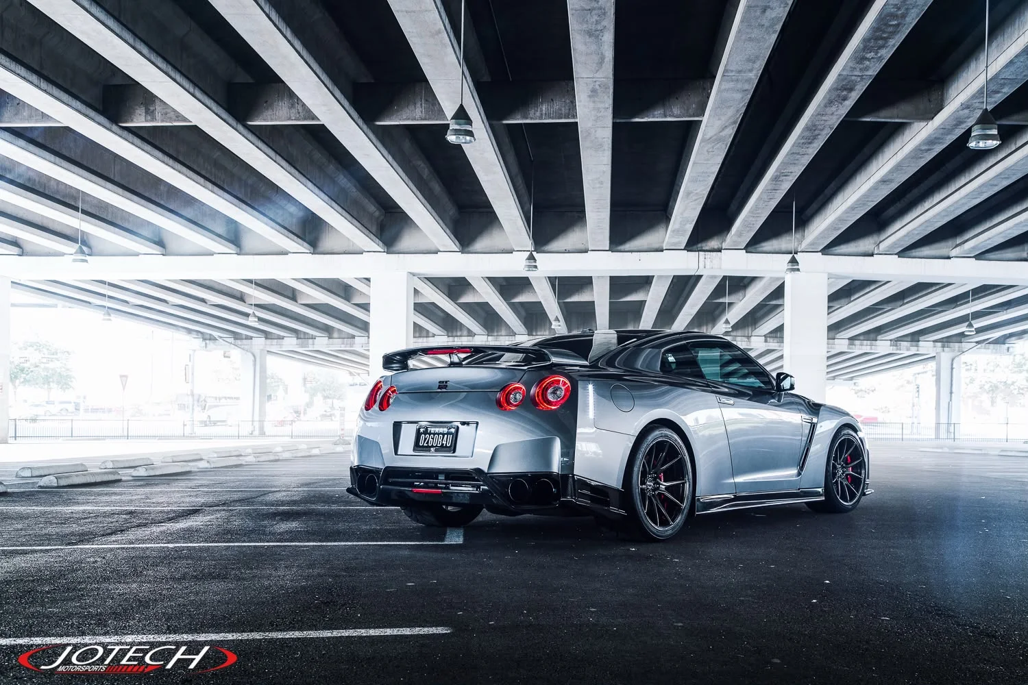 A silver Nissan GT-R with aftermarket wheels and a rear spoiler parked under a concrete overpass.