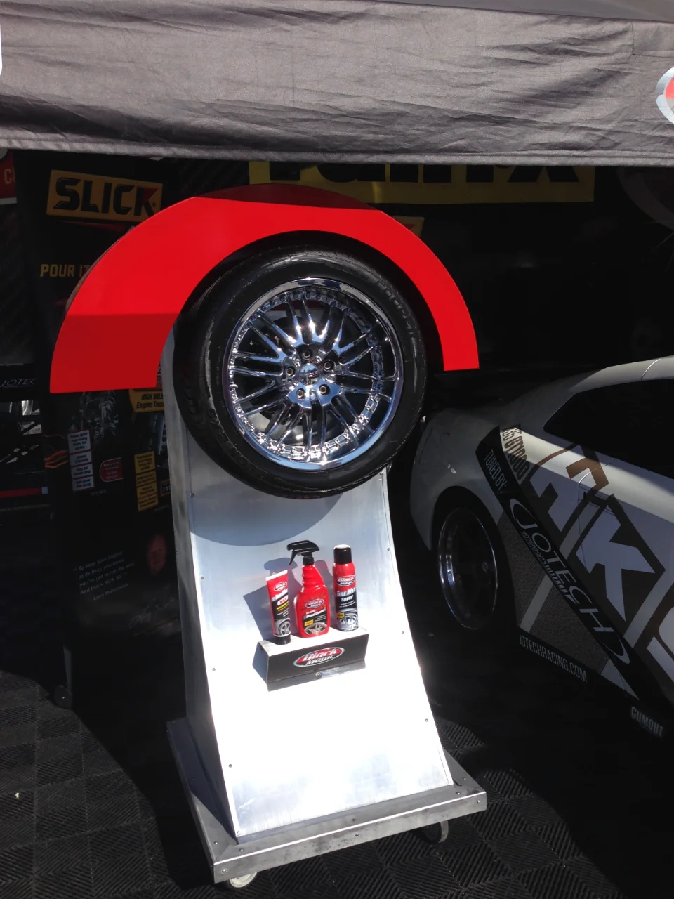A chrome wheel and tire display with car care products at a Jotech Motorsports event booth, with a Jotech branded car in the background.