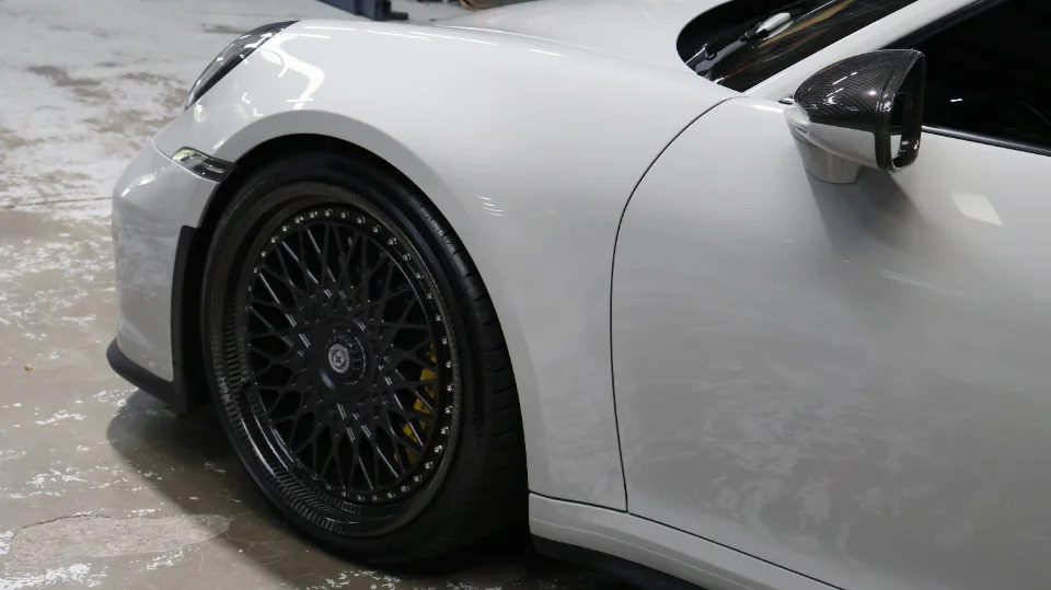 A white Porsche 911 with black HRE wheels and yellow brake calipers, featuring a carbon fiber side mirror.