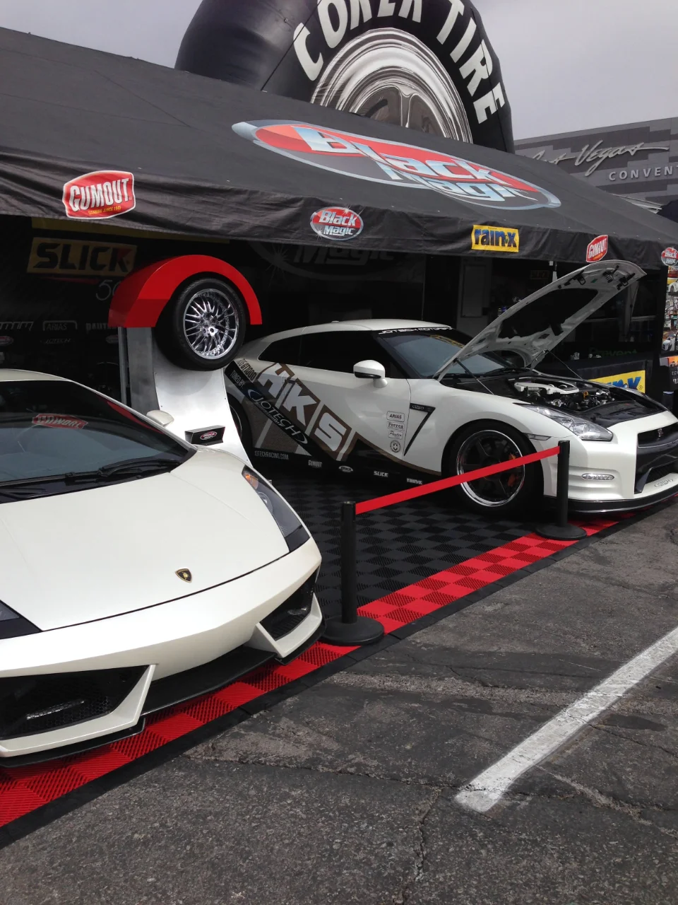 White Lamborghini and HKS-liveried Nissan GT-R on display at an outdoor event booth under a Coren Tire tent.