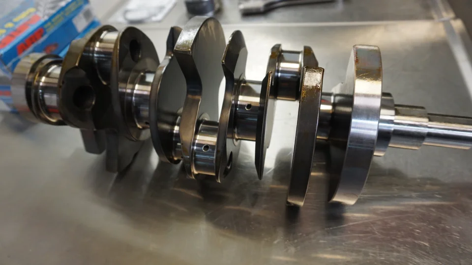A close-up view of a high-performance crankshaft resting on a metallic workbench in a shop setting.