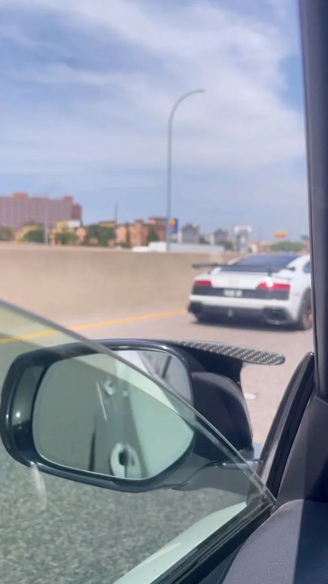 A white Audi R8 with a large carbon fiber wing drives on a highway, viewed from the passenger side of another car.