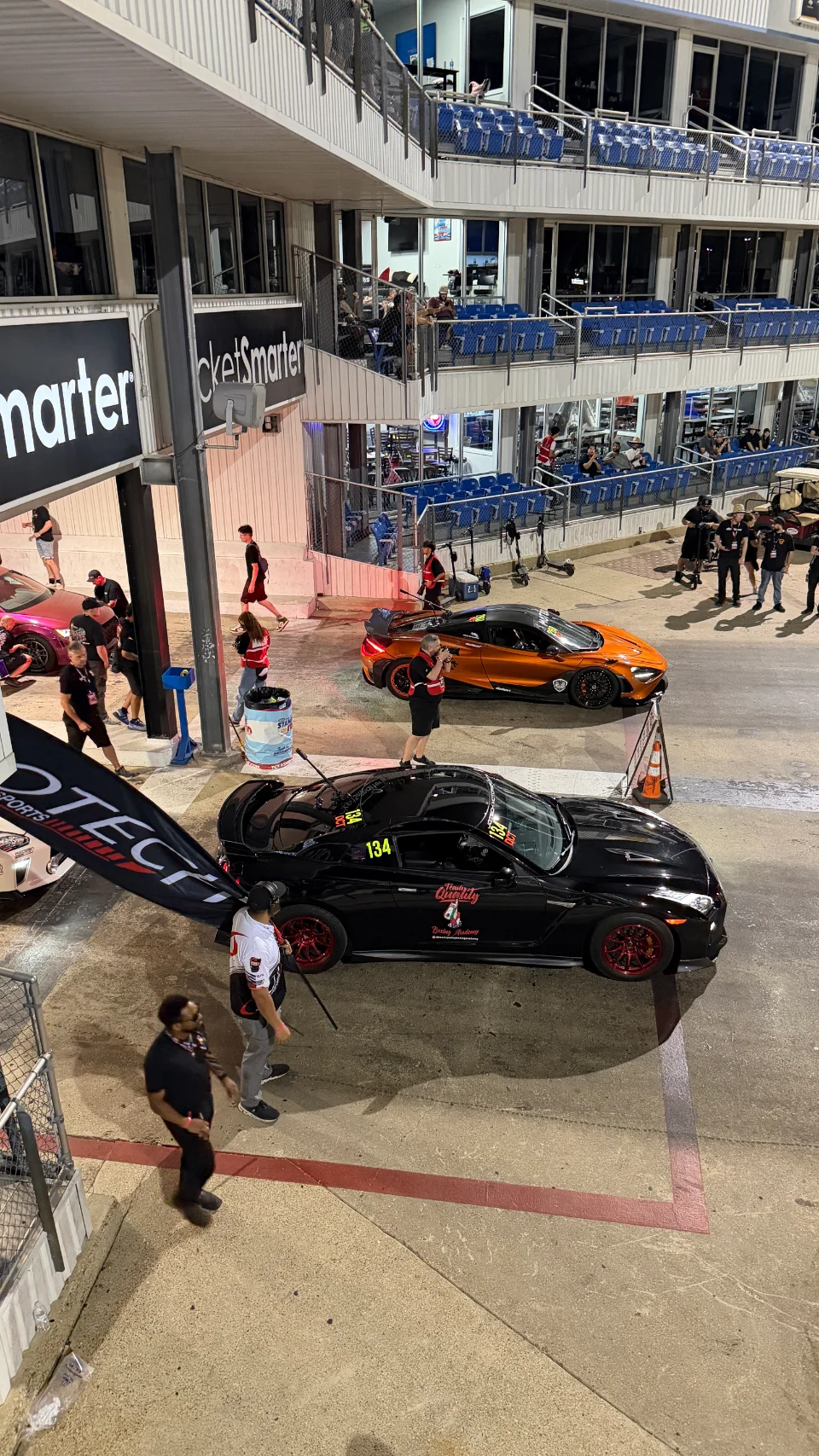 An overhead view of a black Nissan GT-R and an orange McLaren in the staging lanes at a drag strip at night.