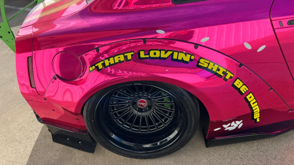 Close-up of a pink chrome wrapped Nissan GT-R with widebody fenders, custom black wheels, and a unique decal.