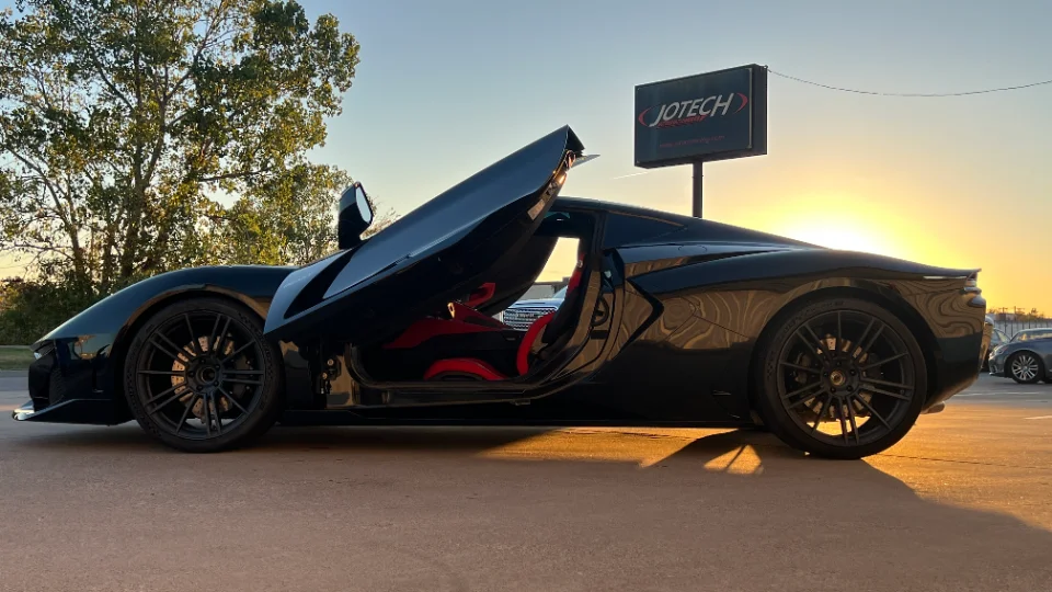 Black McLaren supercar with butterfly doors open, parked outside Jotech Motorsports at sunset, showcasing its sleek design.