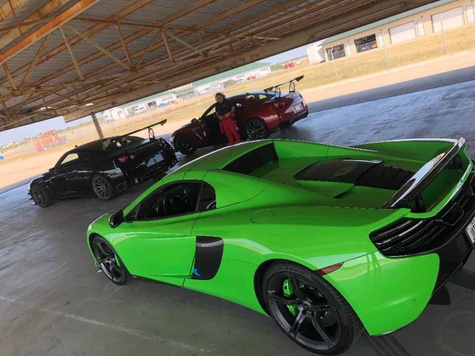 A bright green McLaren and two Nissan GT-Rs parked under a covered area at a track day event.