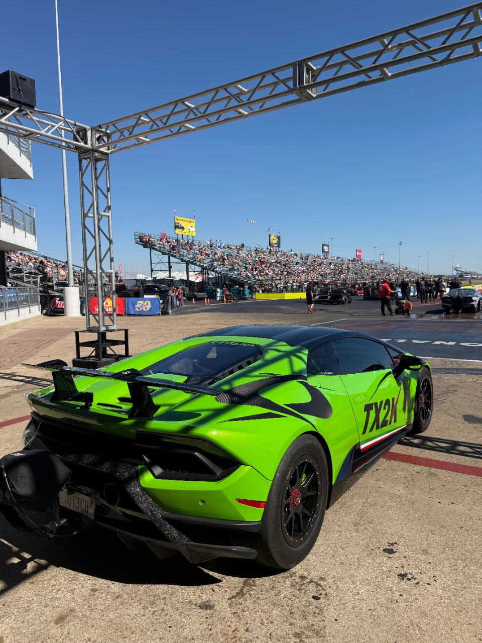 A bright green Lamborghini Huracan with a large rear wing and drag tires at a drag strip during TX2K, with a crowd in the background.