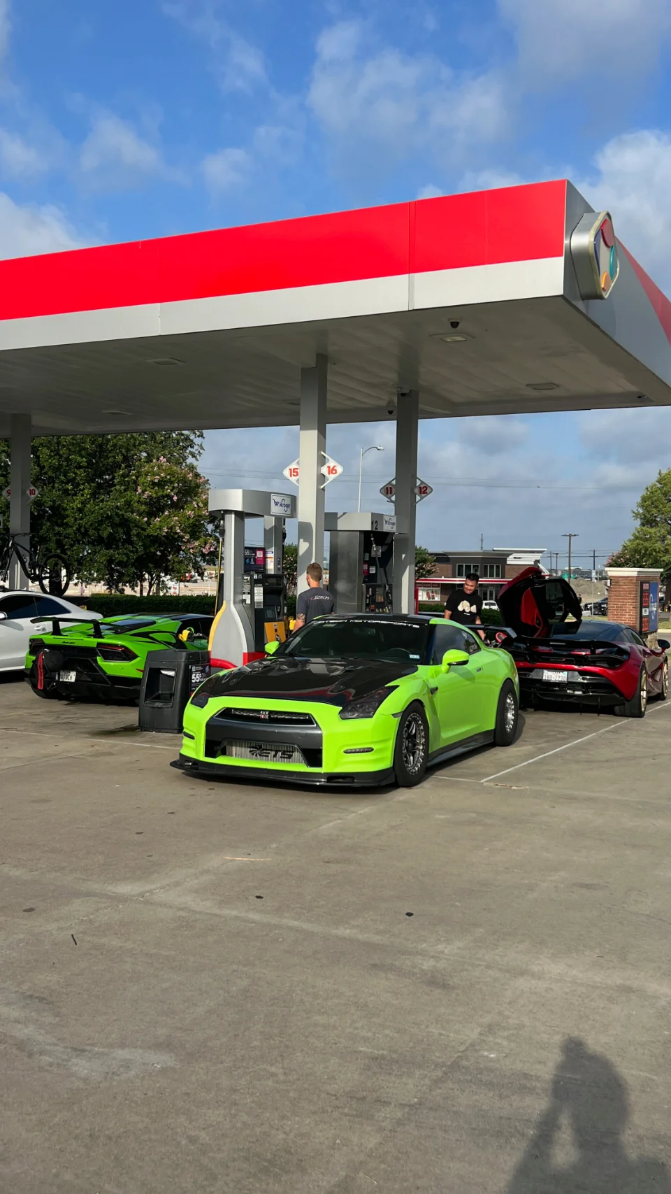 A group of high-performance cars, including a neon green Nissan GT-R, a green Lamborghini, and a red McLaren, at a gas station.