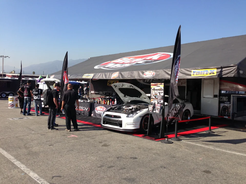 A white Nissan GT-R with its hood open on display under a branded tent at an outdoor event, with people gathered around.