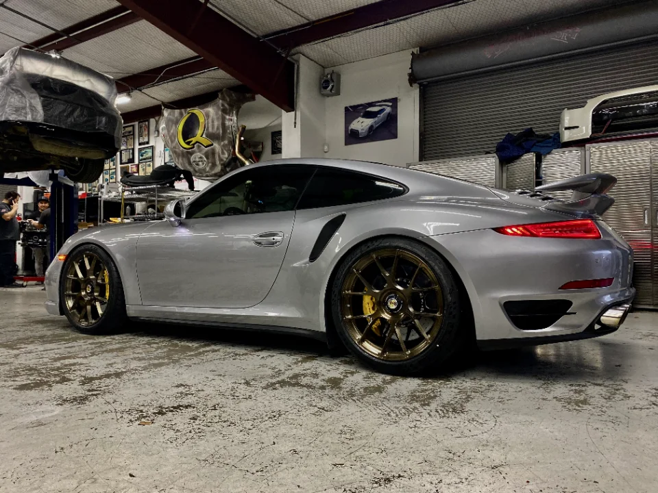 A silver Porsche 911 with custom bronze wheels and yellow calipers parked in a performance tuning shop.
