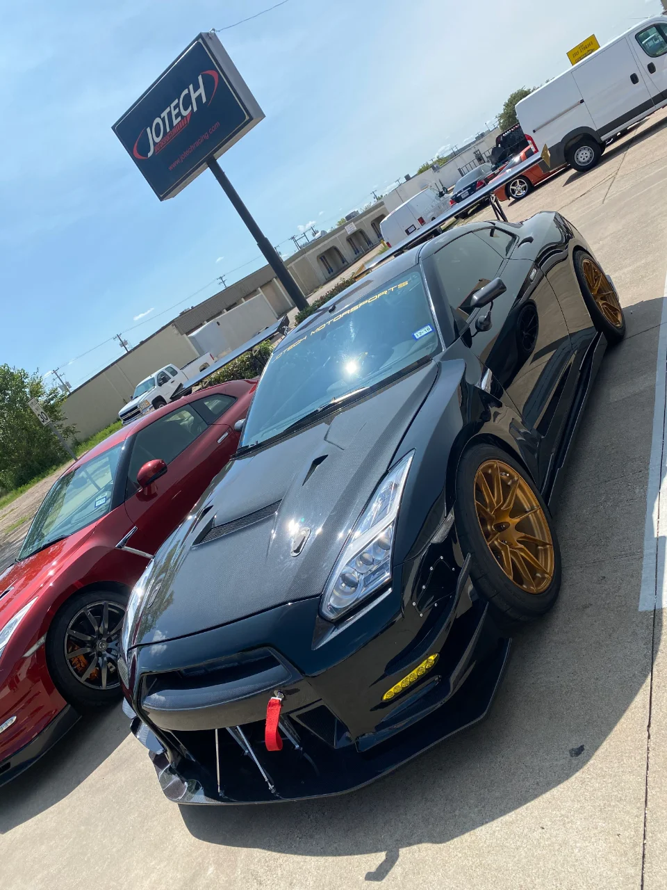 Two Nissan GT-Rs, one black with carbon fiber aero and gold wheels, and one red, parked outside Jotech Motorsports.