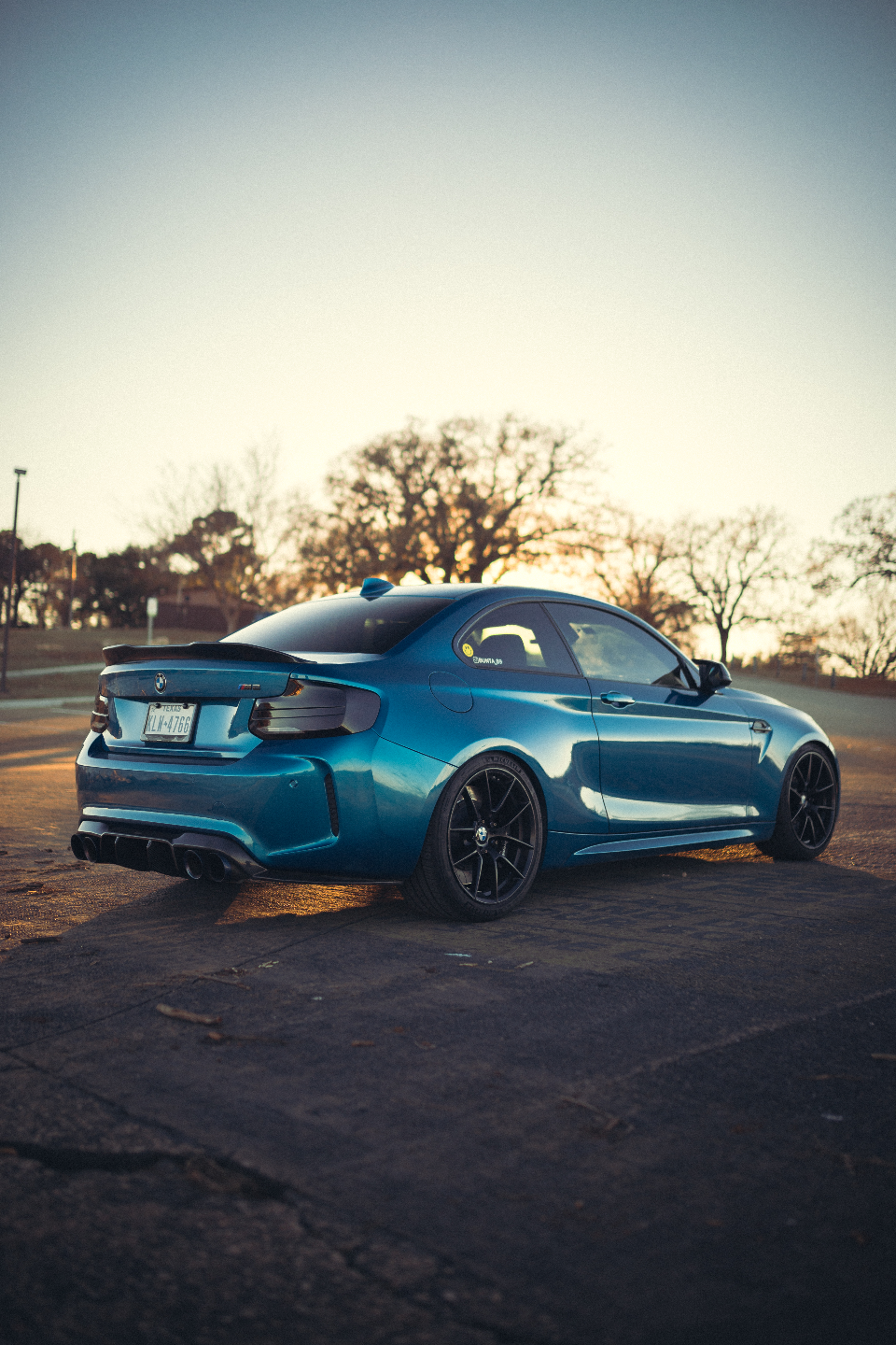 Rear three-quarter view of a blue BMW M2 G87 with aftermarket wheels, rear spoiler, and quad exhaust tips at sunset.