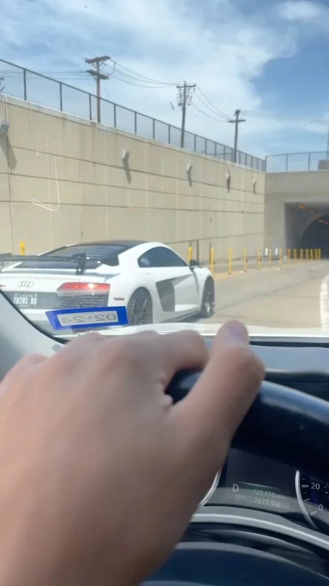 A white Audi R8 with a large rear wing driving on a street, viewed from the perspective of another car's driver.