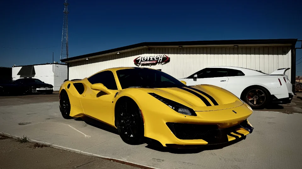 A yellow Ferrari 488 Pista with black racing stripes and a white Nissan GT-R parked outside the Jotech Motorsports facility.