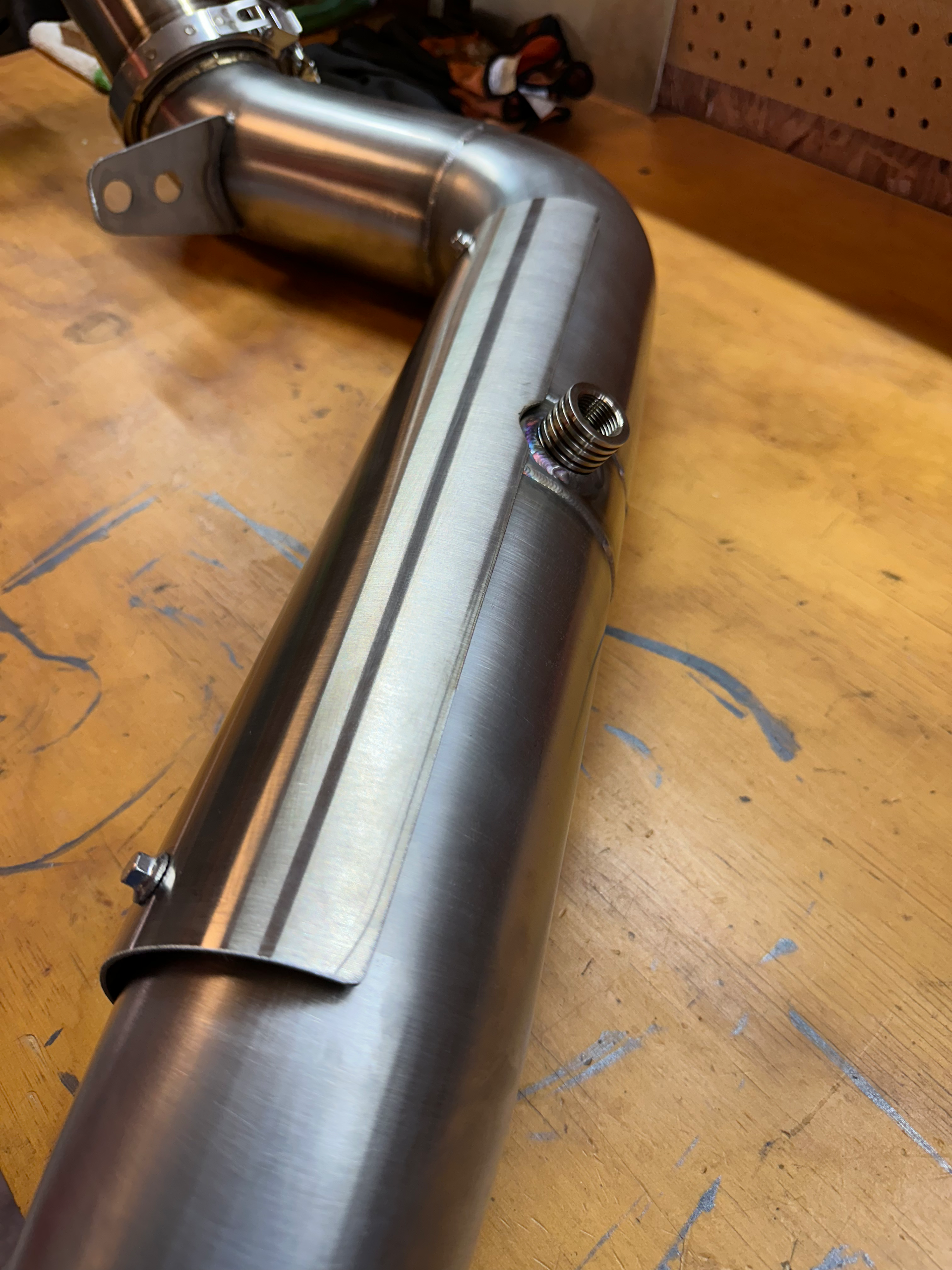 Close-up of a custom fabricated stainless steel exhaust pipe with an O2 sensor bung on a workbench, showcasing the weld quality.