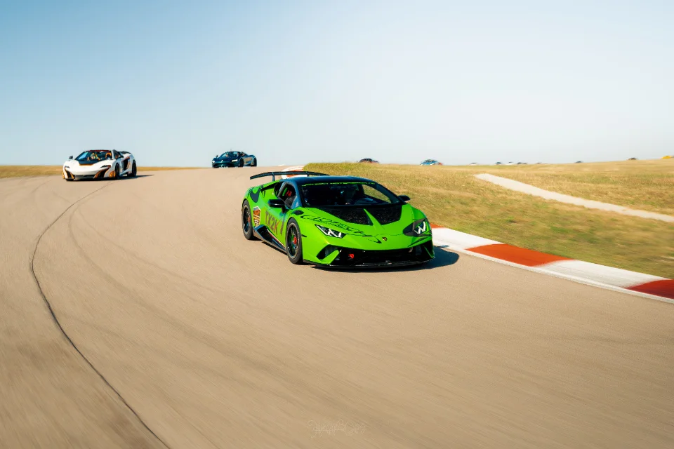 A vibrant green Lamborghini Huracan leads a white McLaren P1 and other supercars on a race track under a clear sky.