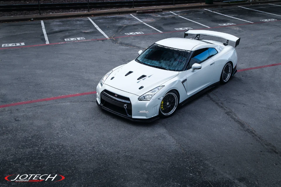 White Nissan GT-R with aftermarket wheels and a large rear wing parked in a lot, showcasing exterior modifications.