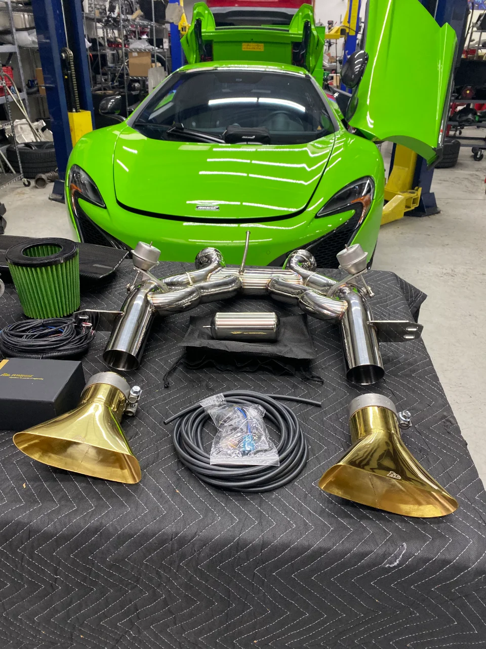 A bright green McLaren supercar in a shop with an aftermarket exhaust system and air filters laid out on a table.