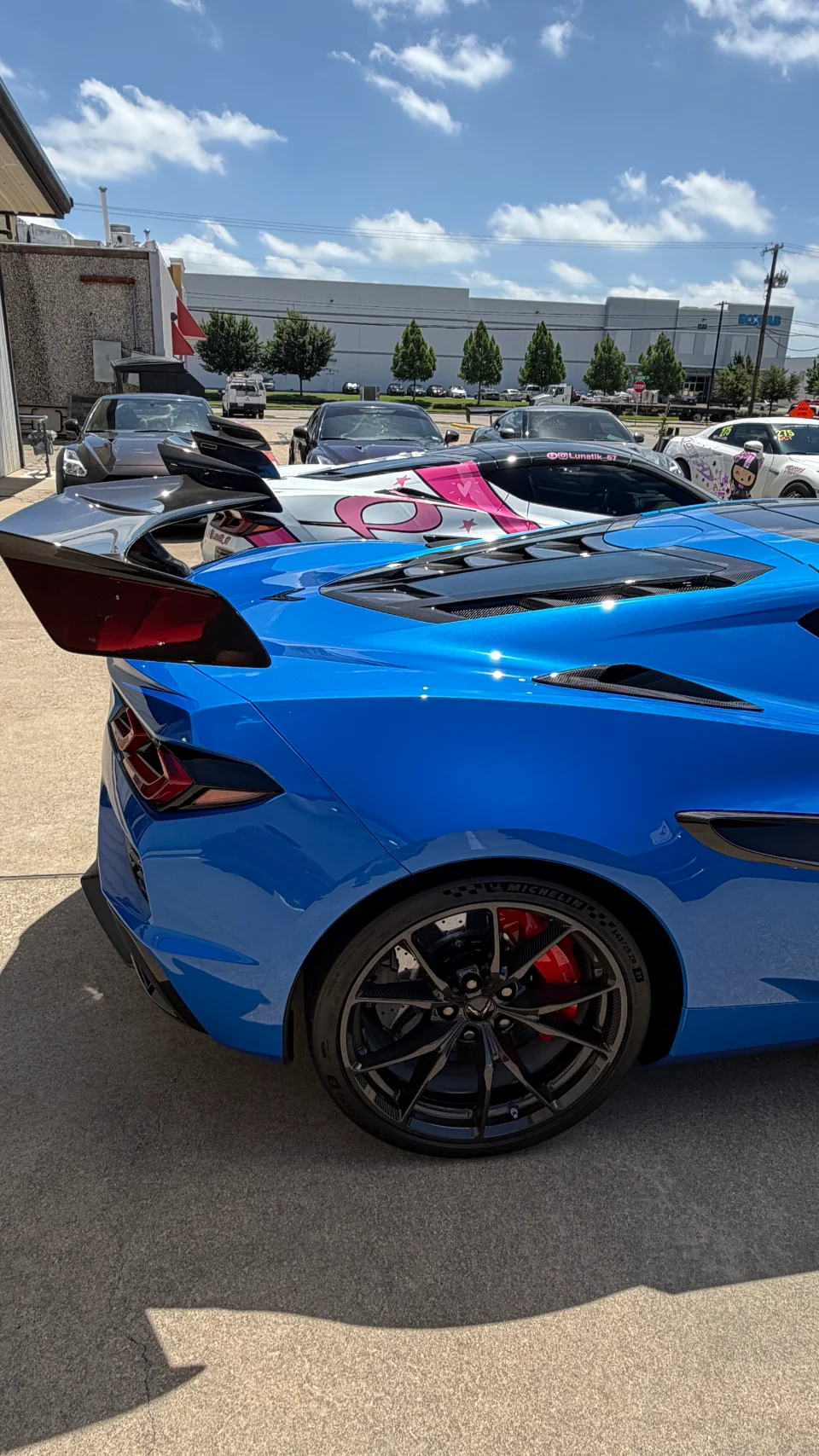 A vibrant blue C8 Corvette with a large carbon fiber rear wing and custom wheels parked at a car meet.