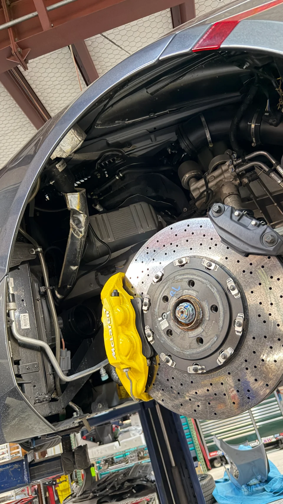 Close-up of a McLaren's yellow brake caliper and drilled rotor on a lift in a performance shop.