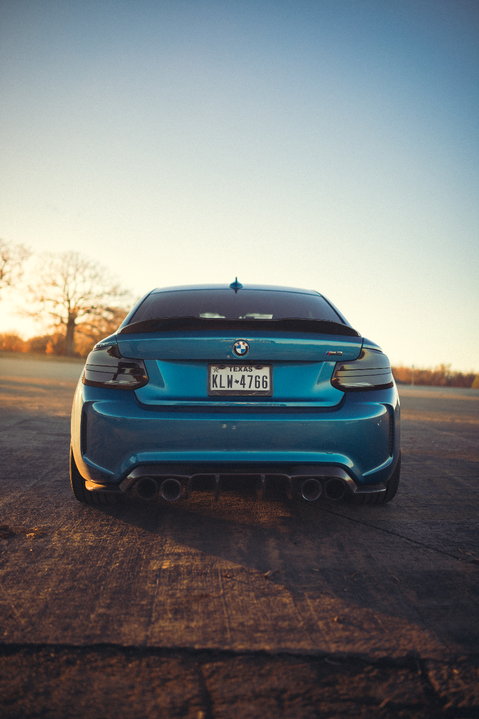 Rear view of a blue BMW M2 with quad exhaust and diffuser at sunset.