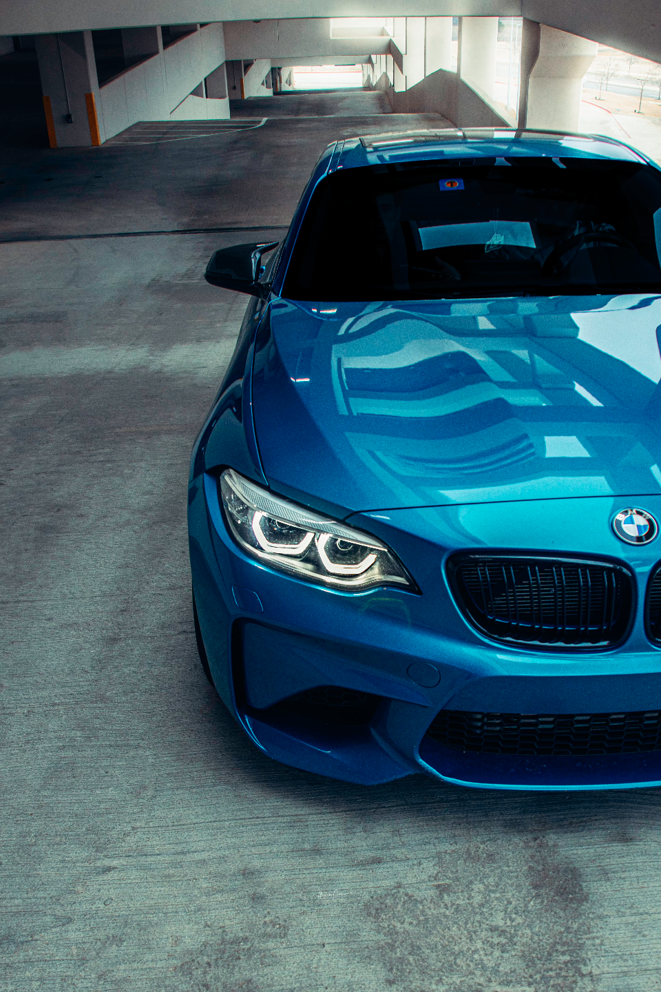 A vibrant blue BMW M2 parked in a concrete parking garage, showcasing its front end and illuminated headlights.
