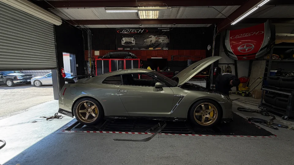 A grey Nissan GT-R strapped to a dyno at Jotech Motorsports, with the hood open and a technician working on the engine.