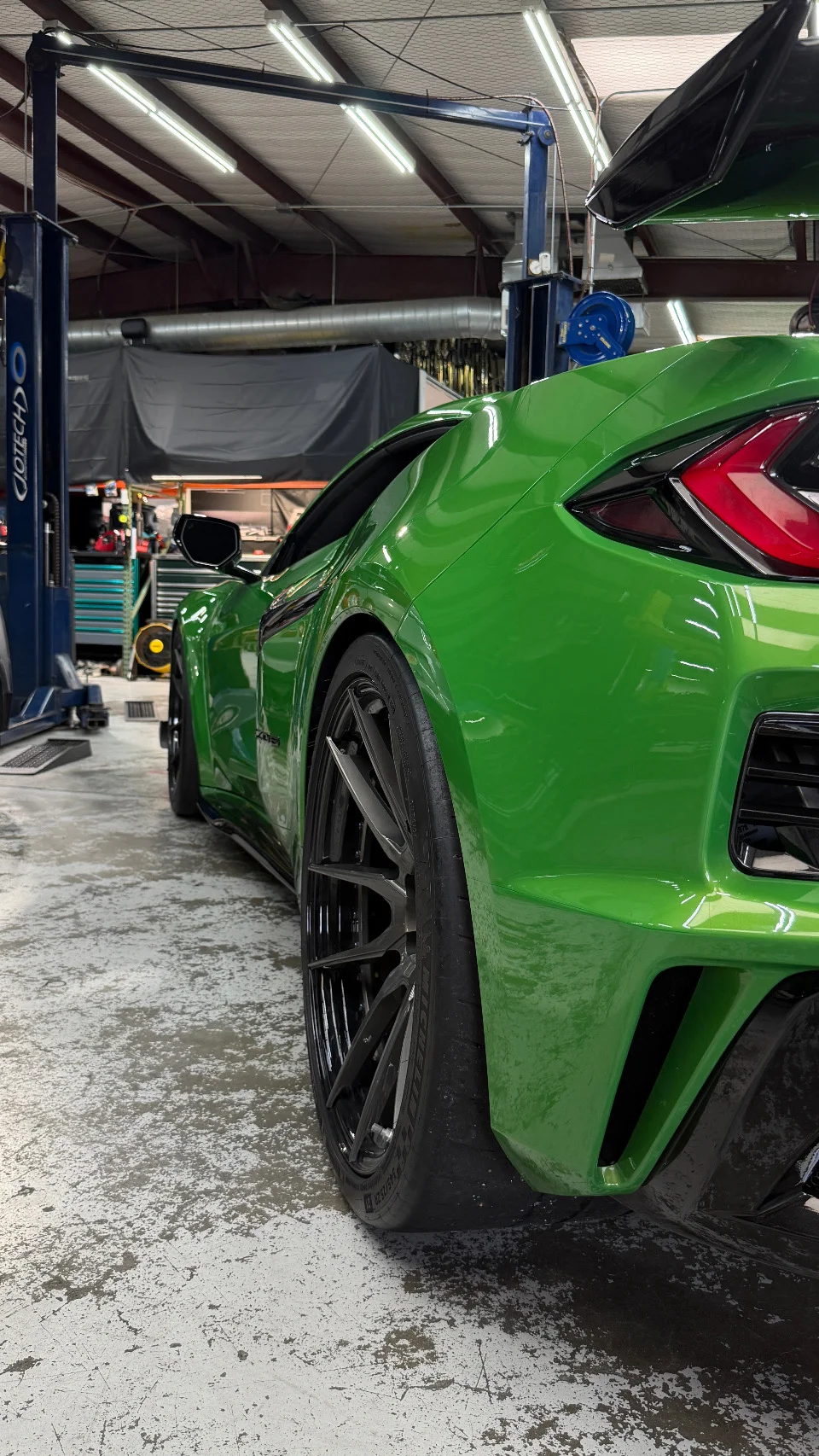 A bright green Corvette C8 on a lift inside a performance tuning shop, showcasing its aftermarket wheels and rear styling.