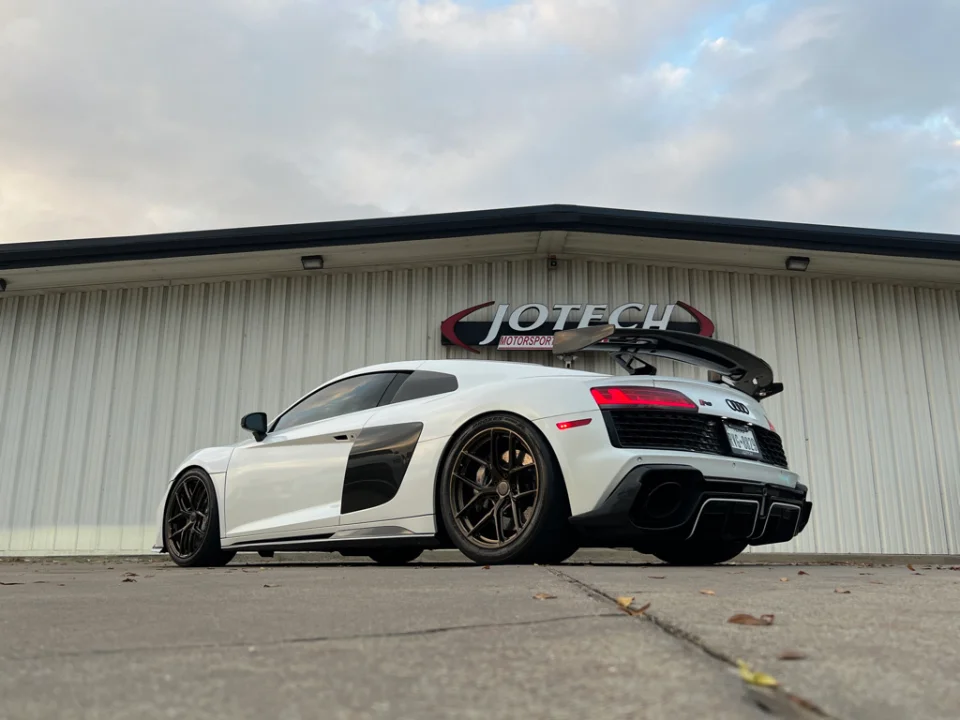 White Audi R8 with aftermarket wheels and large carbon fiber wing parked in front of Jotech Motorsports shop.