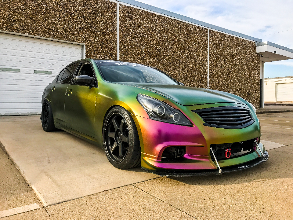 A custom chameleon-wrapped Infiniti G37 sedan with aftermarket wheels and a front splitter parked in front of a building.