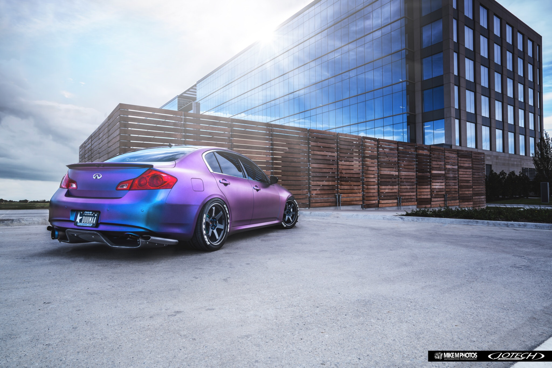 A purple and blue chameleon-wrapped Infiniti G37 sedan with aftermarket wheels parked in front of a modern glass building.