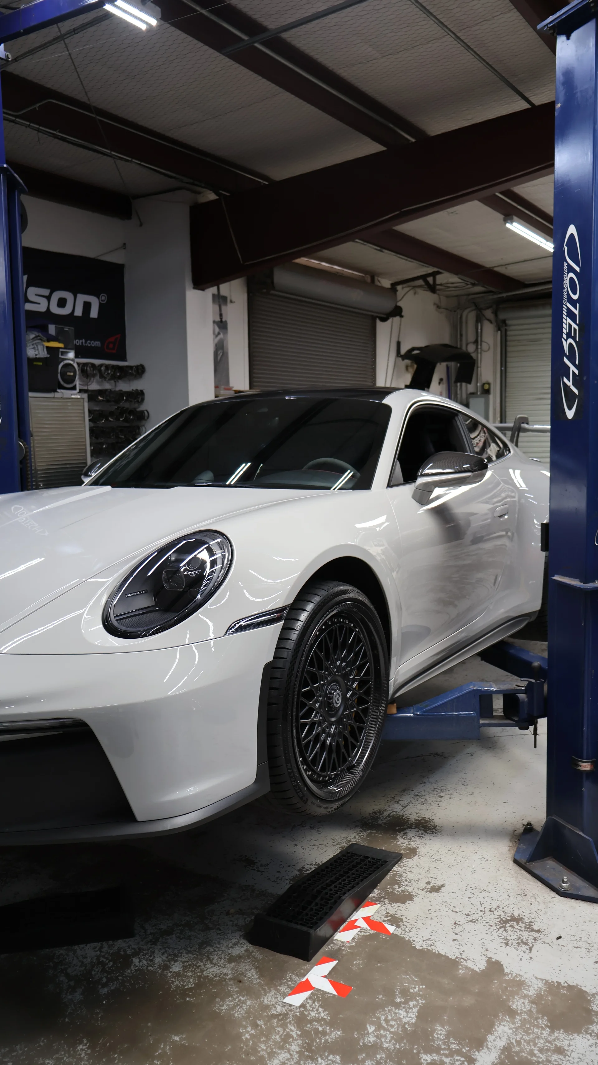 A white Porsche 911 on a hydraulic lift inside the Jotech Motorsports shop, ready for service or modifications.